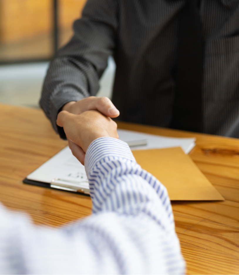Two business professionals shaking hands across a conference table with documents and a folder, symbolizing a commercial lease agreement or successful contract negotiation.